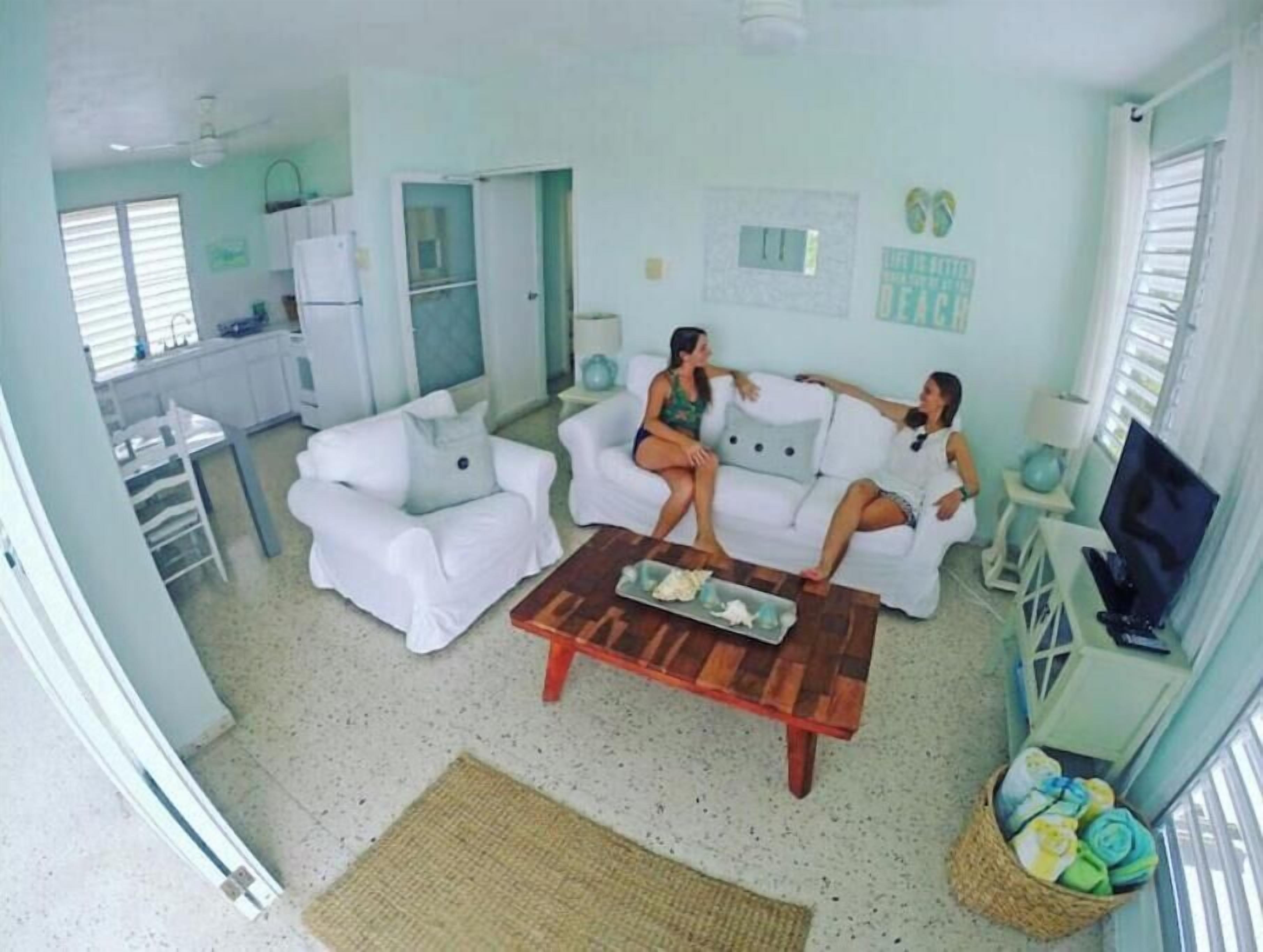 Un saló amb sofàs blancs, una taula de centre de fusta i una televisió. Hi ha una zona de cuina amb armaris blancs i un frigorífic.