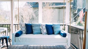 Un canapé blanc pour l’extérieur, agrémenté de coussins bleus et à motifs, est installé sur une terrasse en bois, d’où l’on peut admirer un jardin et une maison.