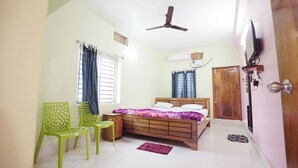 A bedroom with a bed, two chairs, a ceiling fan, a window with curtains, and a door. - Sadhu Inn (Puri)