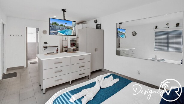 A modern bedroom with a large bed, a TV mounted on the wall, a wardrobe, and a bathroom visible through an open door.