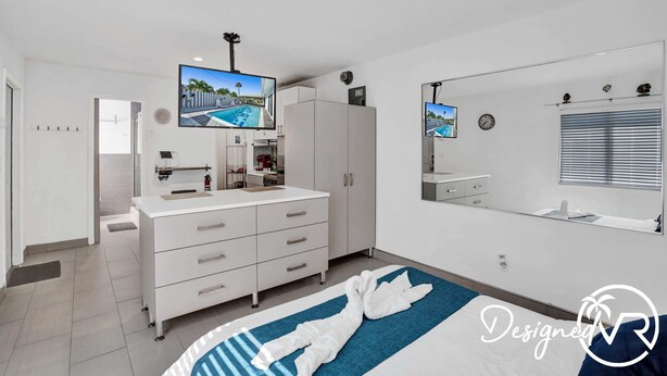 A modern bedroom with a large bed, a TV mounted on the wall, a wardrobe, and a bathroom visible through an open door.