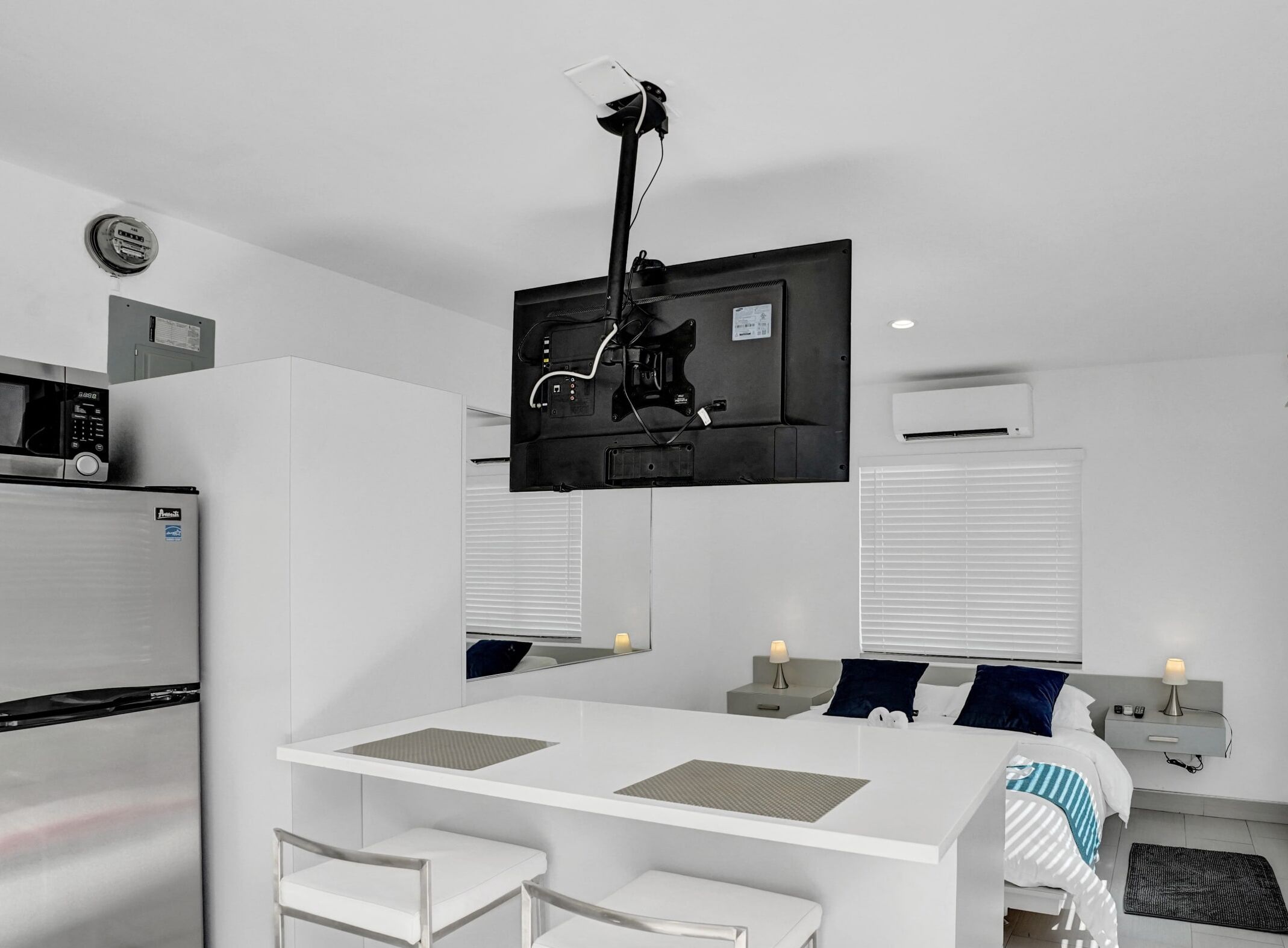 A modern hotel room with a flat-screen TV mounted on the wall, a kitchenette with a fridge and microwave, a dining area with a table and chairs, and a bed with a nightstand and lamp.