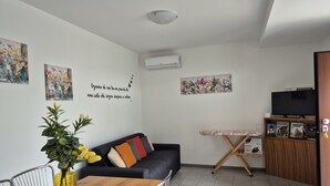 Ein modernes Wohnzimmer mit einer Couch, einem Esstisch und Stühlen. An den Wänden befinden sich Kunstwerke, eine Zimmerpflanze und ein Fernseher auf einem Standfuß.