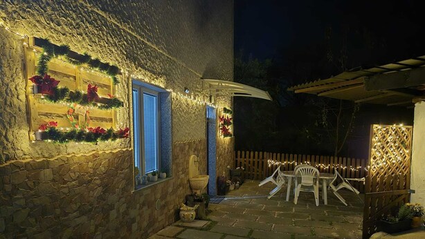 Ein Steinhaus mit einer Holztür und Fenstern, die mit Lichtern und Weihnachtssternen geschmückt sind. Vor dem Haus befindet sich eine Terrasse mit Tisch und Stühlen, im Hintergrund ein Holzzaun.