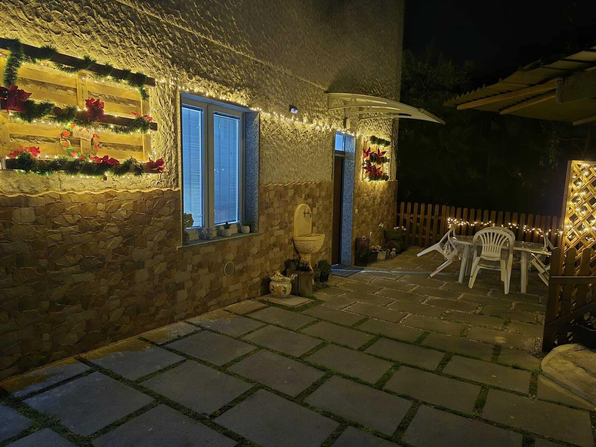 A stone-walled patio with a stone floor, a wooden table and chairs, and a pergola with string lights.
