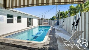 Una piscina con solárium de baldosas, sillas de descanso y una zona cubierta con toldo a rayas.