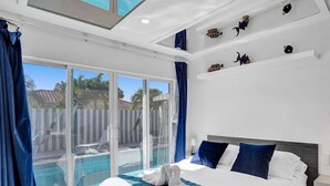 Habitación de hotel con cama, mesita de noche, lámpara y vista a una piscina a través de amplios ventanales.