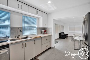 A modern kitchen with white cabinets, a stainless steel sink, and a coffee maker. There is a dining area with a table and chairs, and a black leather sofa. - Unit C Sea&i Private House Near Deerfield Beach (Deerfield Beach)