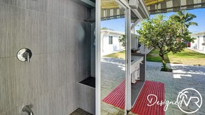 Une cabine de douche moderne dotée d’un panneau de commande et offrant une vue sur un espace extérieur abrité, avec un arbre et un bâtiment en arrière-plan.