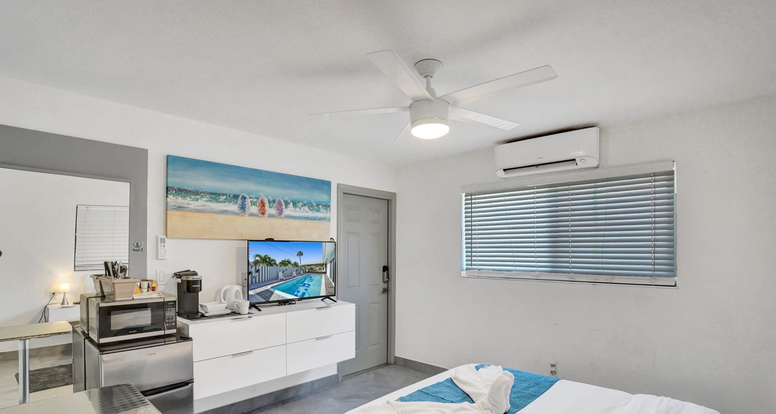 A hotel room with a bed, a desk with a microwave, a coffee maker, and a TV displaying a beach scene.