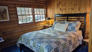 Dormitorio de cabaña de madera con cama, mesita de noche, lámpara y ventana con vista a un vehículo fuera.