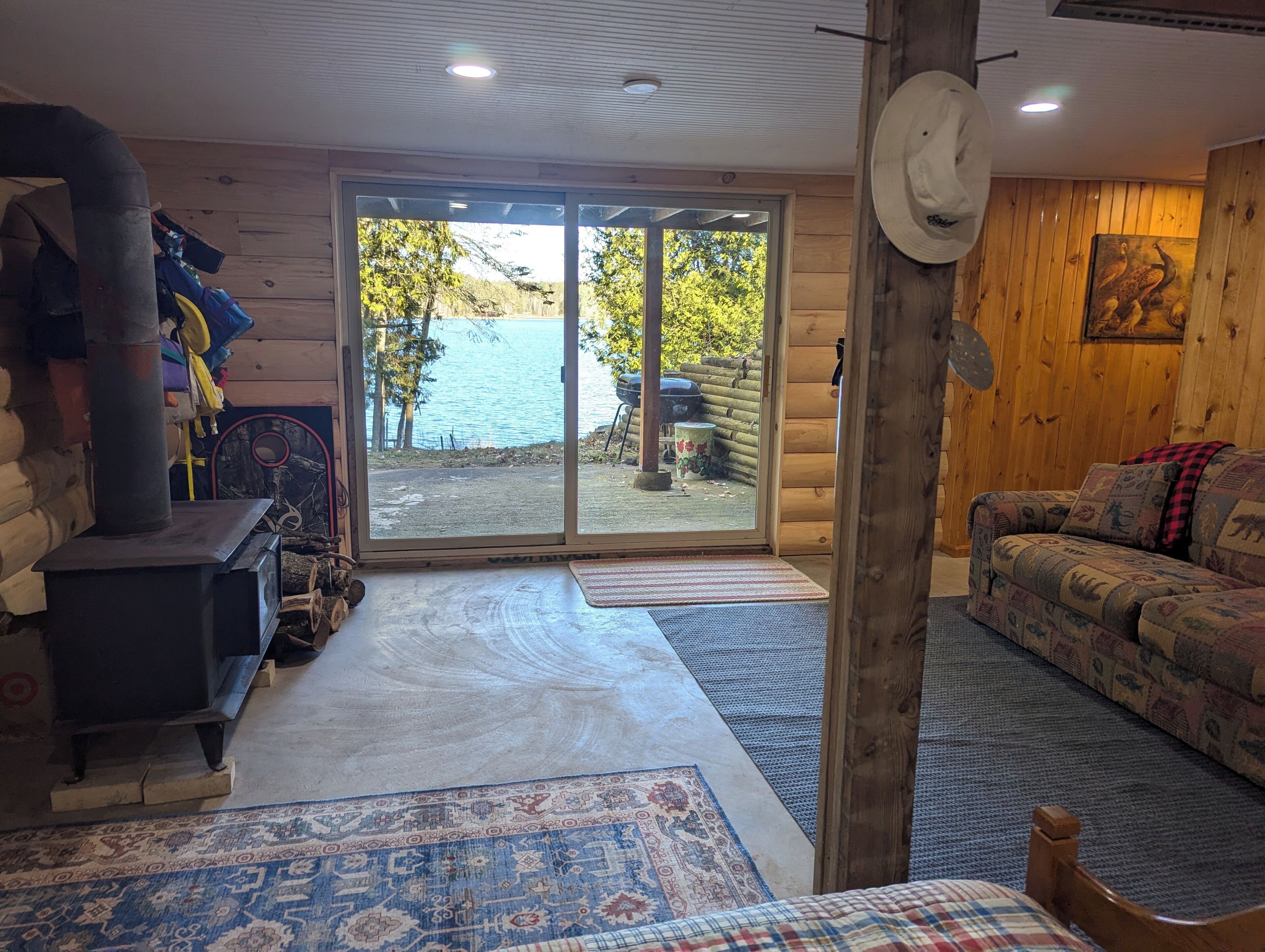 A log cabin interior with a wood-burning stove, a sofa, and a view of a lake through large glass doors.
