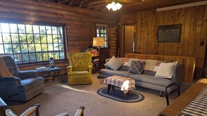 Un acogedor interior de cabaña de madera con un sofá, un sillón y un otoman dispuestos alrededor de una alfombra central.