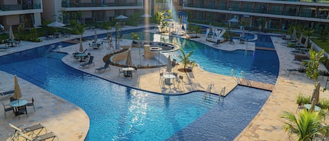 A large outdoor swimming pool surrounded by lounge chairs and umbrellas, with a central water feature and multiple levels of buildings in the background.