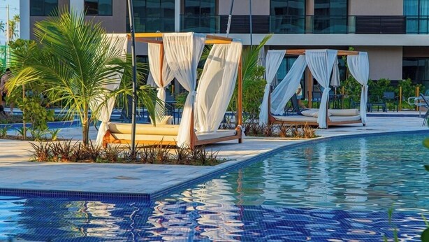 Zona de piscina con dos camas con dosel, palmeras y un edificio moderno al fondo.