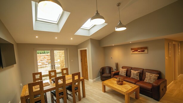 A spacious living area with a dining table, chairs, a sofa, and a coffee table. There are skylights and pendant lights.