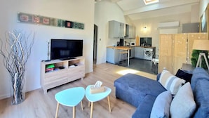 Ein modernes Wohnzimmer mit einem Flachbildfernseher, einer Couch und einem Couchtisch. Im Hintergrund ist eine Küche mit einem Essbereich zu sehen.