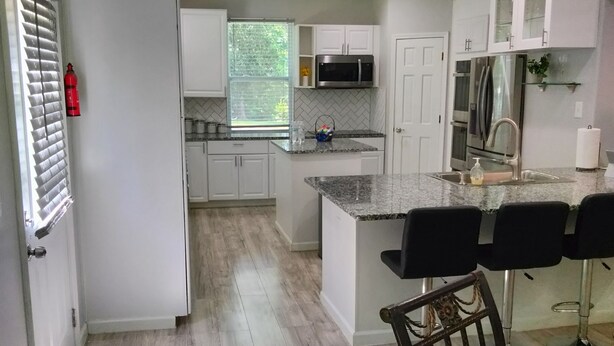 Eine moderne Küche mit weißen Schränken, einer Granit Arbeitsplatte, einem Edelstahlkühlschrank und einer Mikrowelle. Eine Glas Kochinsel mit Barhocker und einem kleinen Spülbecken.