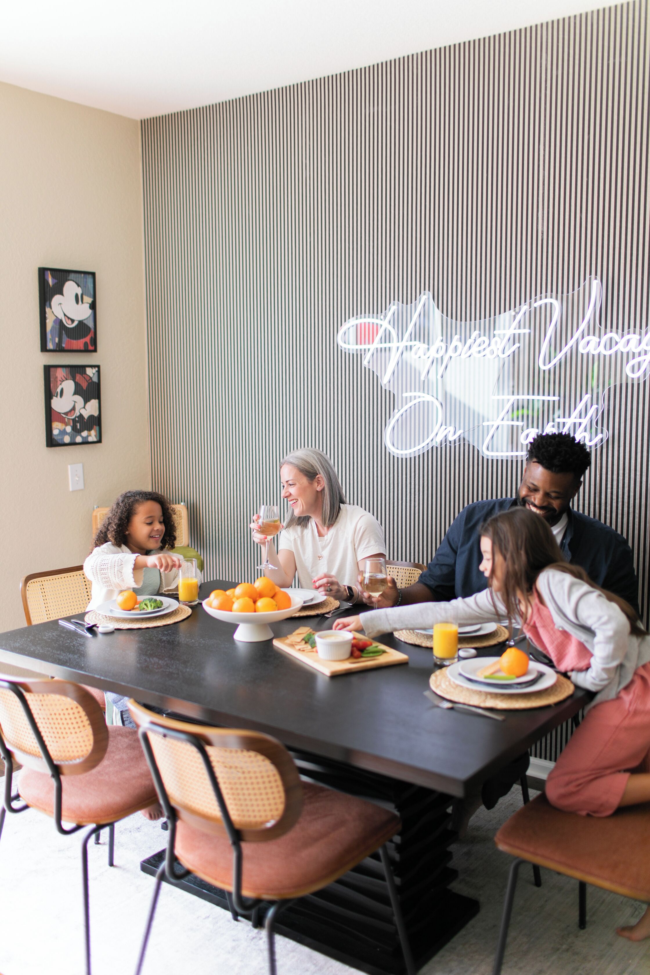 A dining table with people seated around it, plates with food and drinks, and a neon sign on the wall.