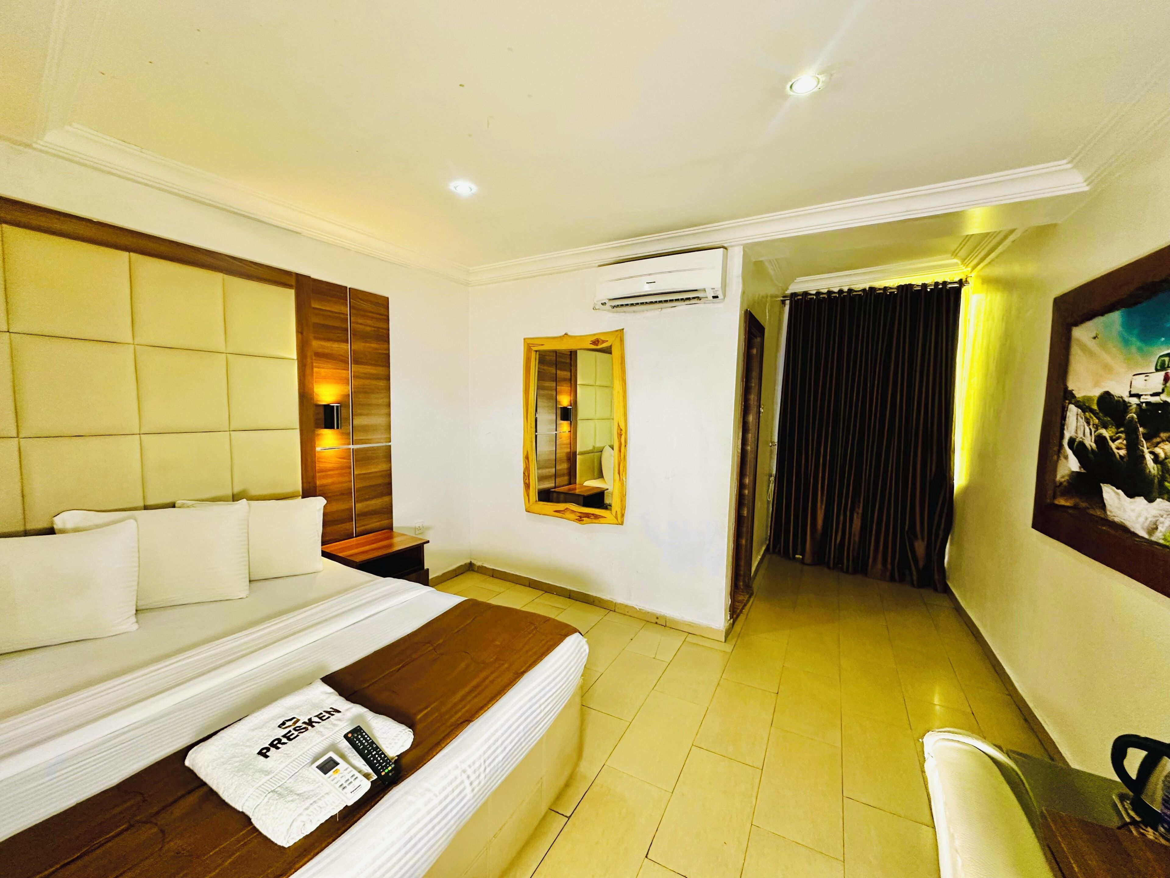 A hotel room with a bed, a bedside table, a mirror, an air conditioning unit, and a painting on the wall.