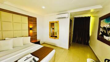 A hotel room with a bed, a bedside table, a mirror, an air conditioning unit, and a painting on the wall.