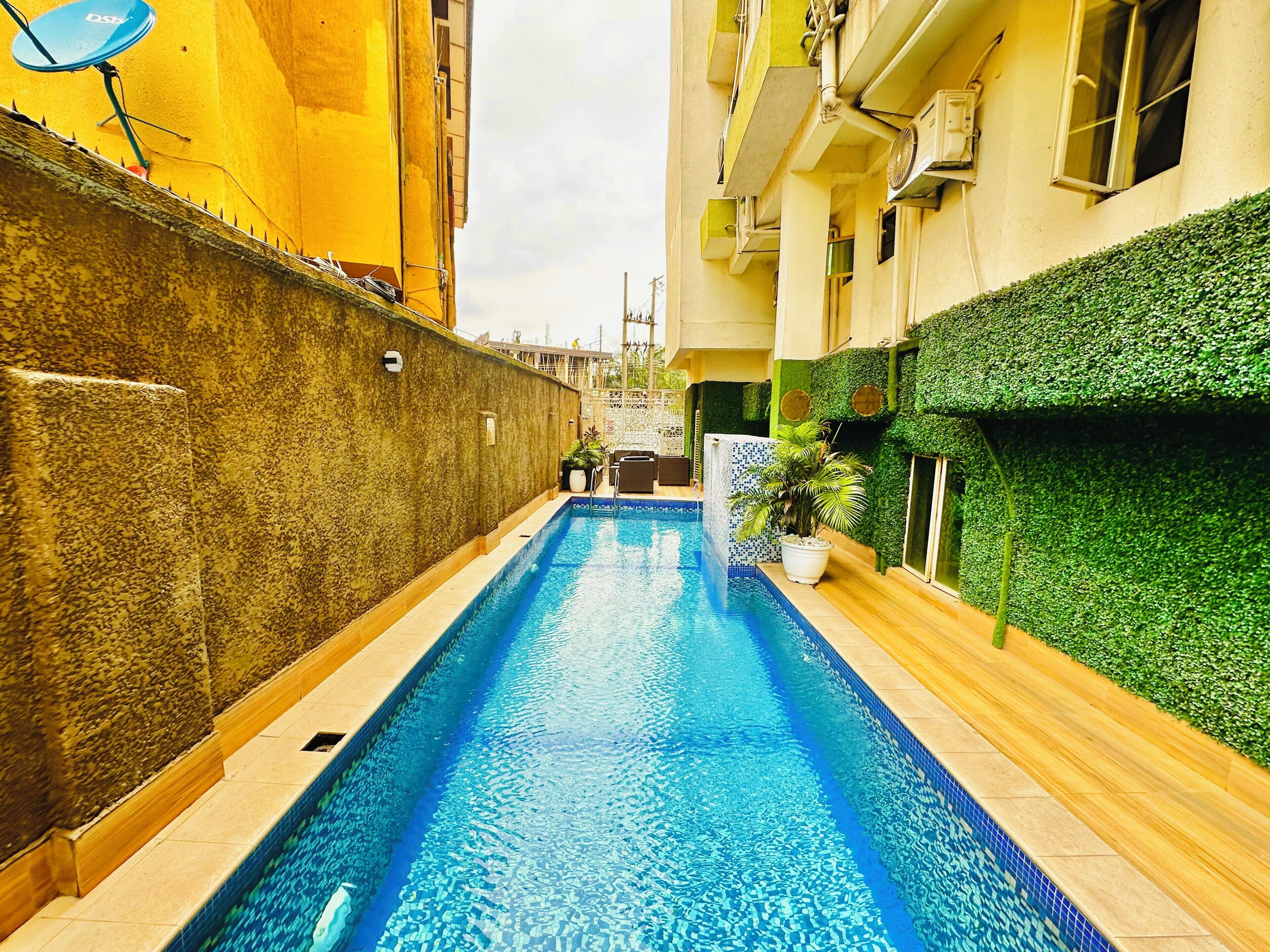 A narrow swimming pool with tiled edges, surrounded by buildings with balconies and a green wall.