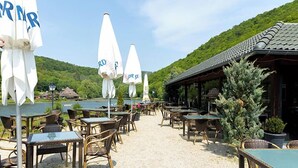 An outdoor seating area with wicker tables and chairs, white umbrellas, and a view of a lake and hills.