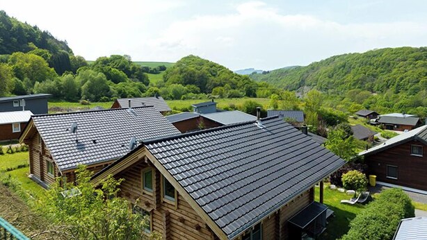 A wooden cabin with a sloped roof, surrounded by lush greenery and hills in the background.