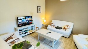 A modern living room with a white sofa, a coffee table, and a TV on a white stand. There is a small side table with a lamp and a picture frame on the wall. - Town house 10 min from Futuroscope (Migné-Auxances)
