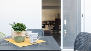 Un coin repas avec une table dressée pour deux personnes, agrémenté d’un pot de fleurs en fibres tressées, d’une tasse à rayures et d’une tasse blanche unie. La table est posée sur un tapis texturé, et deux chaises à dossier ajouré se trouvent à proximité. L’arrière-plan comprend un espace cuisine avec des appareils modernes et une table à manger.