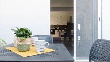 Un comedor con una mesa dispuesta para dos, que incluye una maceta tejida, una taza a rayas y una taza blanca lisa. La mesa está sobre un felpudo texturizado, y hay dos sillas con respaldos perforados. El fondo muestra una cocina con electrodomésticos modernos y una mesa de comedor.