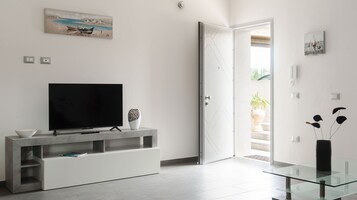 Un salón moderno con un televisor de pantalla plana sobre una consola blanca, una mesa de centro de vidrio y una puerta que da a un espacio exterior.