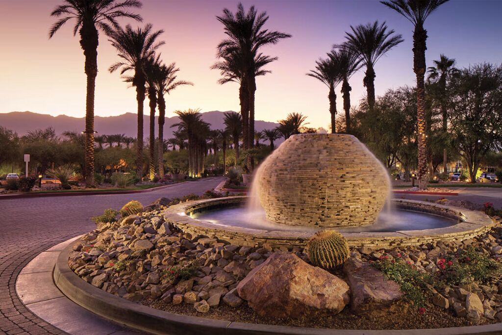A circular stone fountain with water jets, surrounded by a landscaped area with rocks and cacti, palm trees, and a mountainous backdrop.