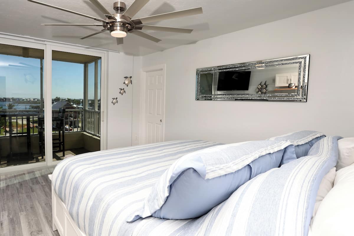 Une chambre avec un grand lit, un ventilateur de plafond, un balcon avec vue et un miroir au mur.