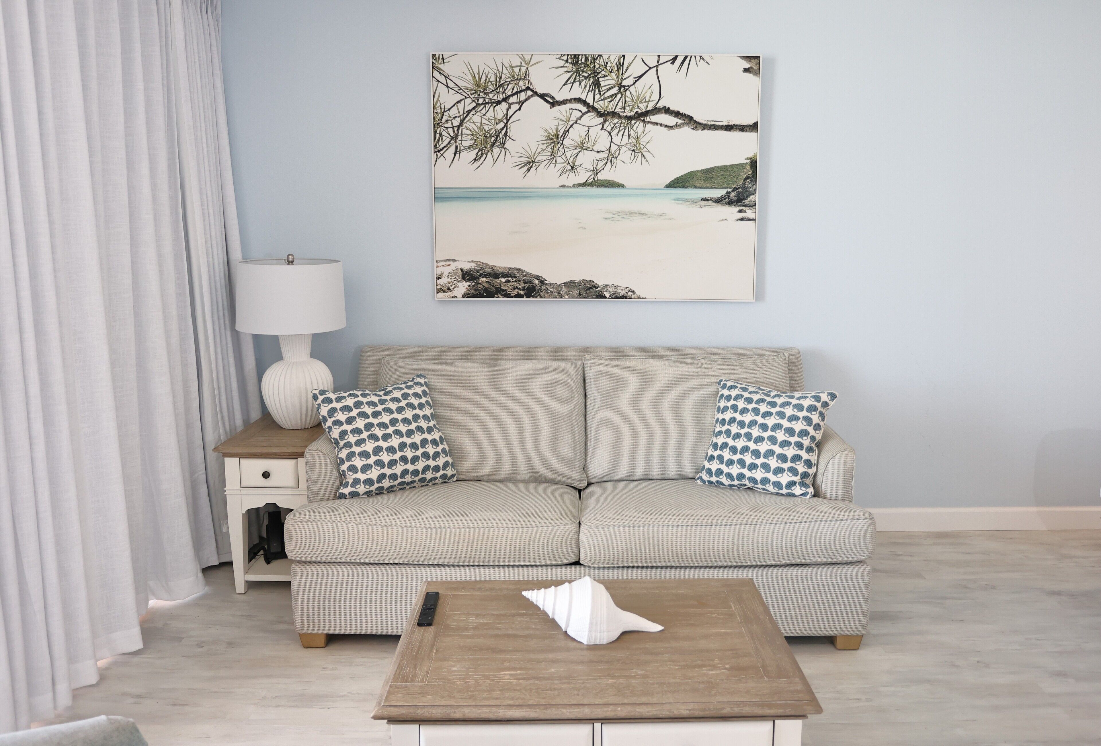 A living room with a beige sofa, patterned pillows, a wooden coffee table, a lamp, and a beach-themed painting on the wall.