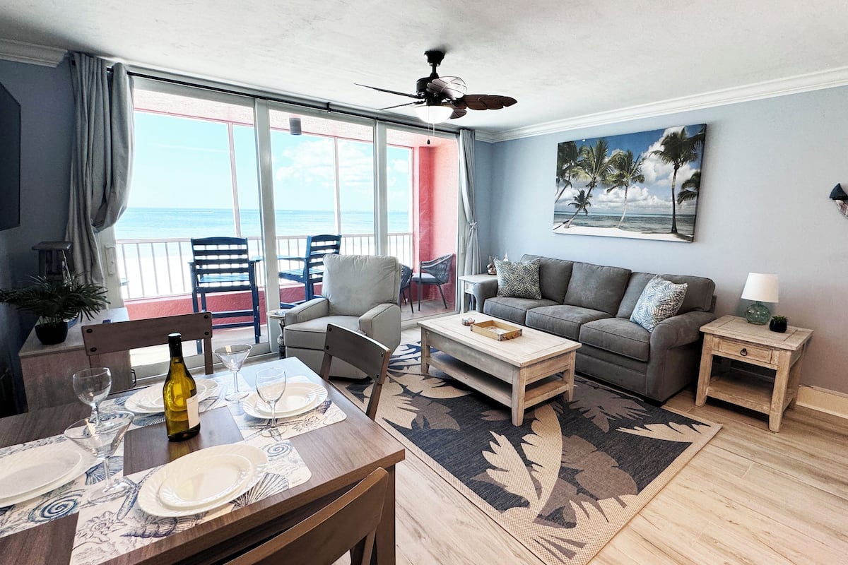 Un salon avec un canapé, une table basse et des fauteuils, un coin repas avec une table dressée pour deux personnes, et un balcon avec des chaises.