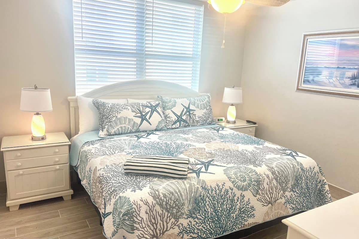 Une chambre à coucher comprenant un lit, deux tables de chevet avec des lampes, un ventilateur de plafond et une fenêtre avec des stores.
