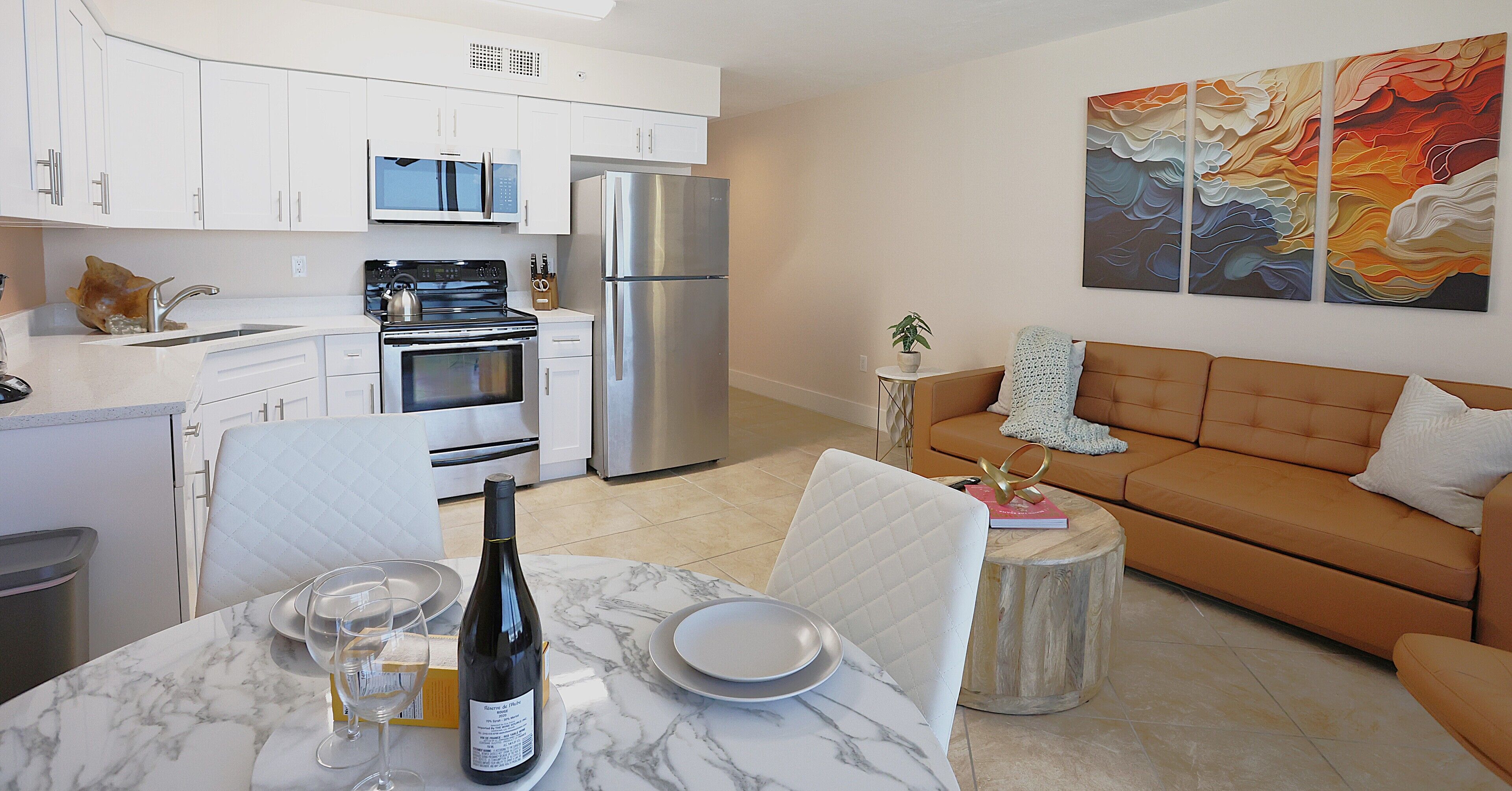 A modern kitchen with white cabinetry, stainless steel appliances, and a marble dining table set with plates, glasses, and a bottle of wine.