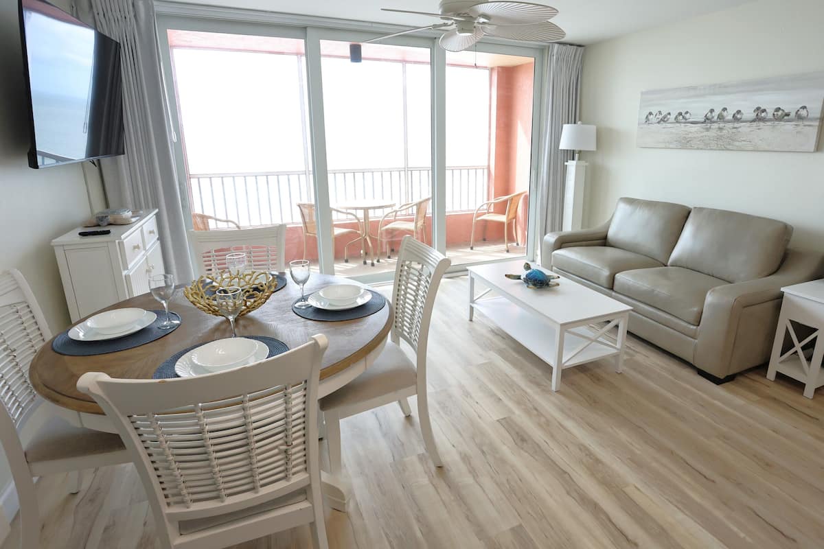 Un salon bien éclairé comprenant un coin repas, un canapé, un téléviseur et un balcon avec des chaises.