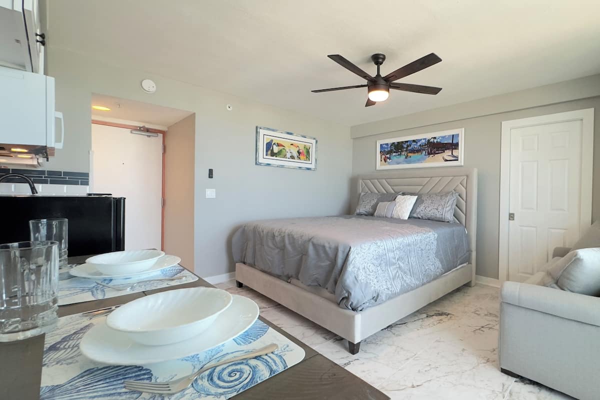 Une chambre à coucher comprenant un lit, une chaise, une table à manger avec des assiettes et des verres, un ventilateur de plafond et deux œuvres d’art murales.