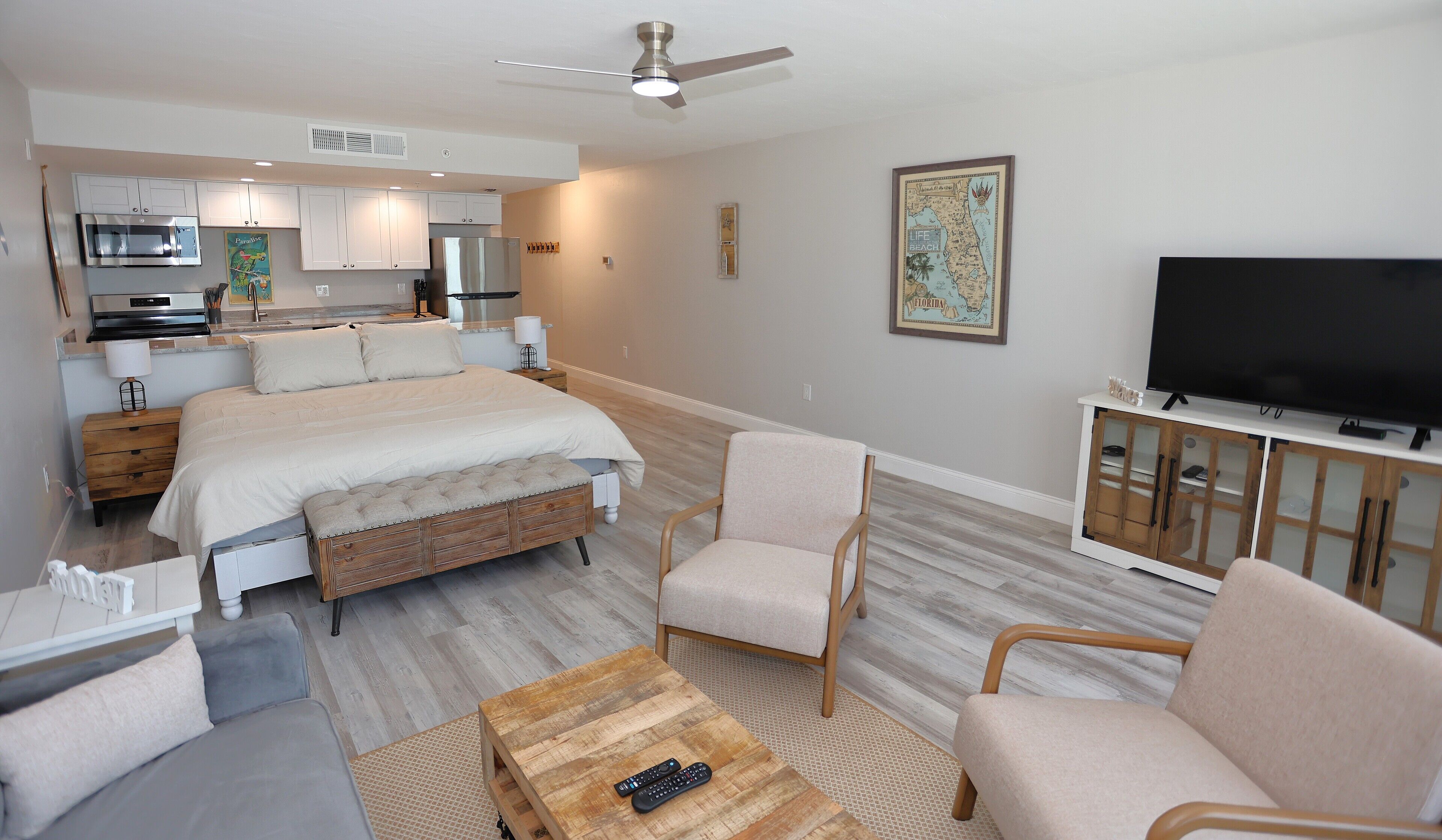 A modern bedroom with a bed, bedside tables, a TV, and a kitchenette in the background.