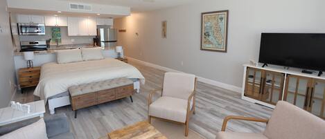 A modern bedroom with a bed, bedside tables, a TV, and a kitchenette in the background.