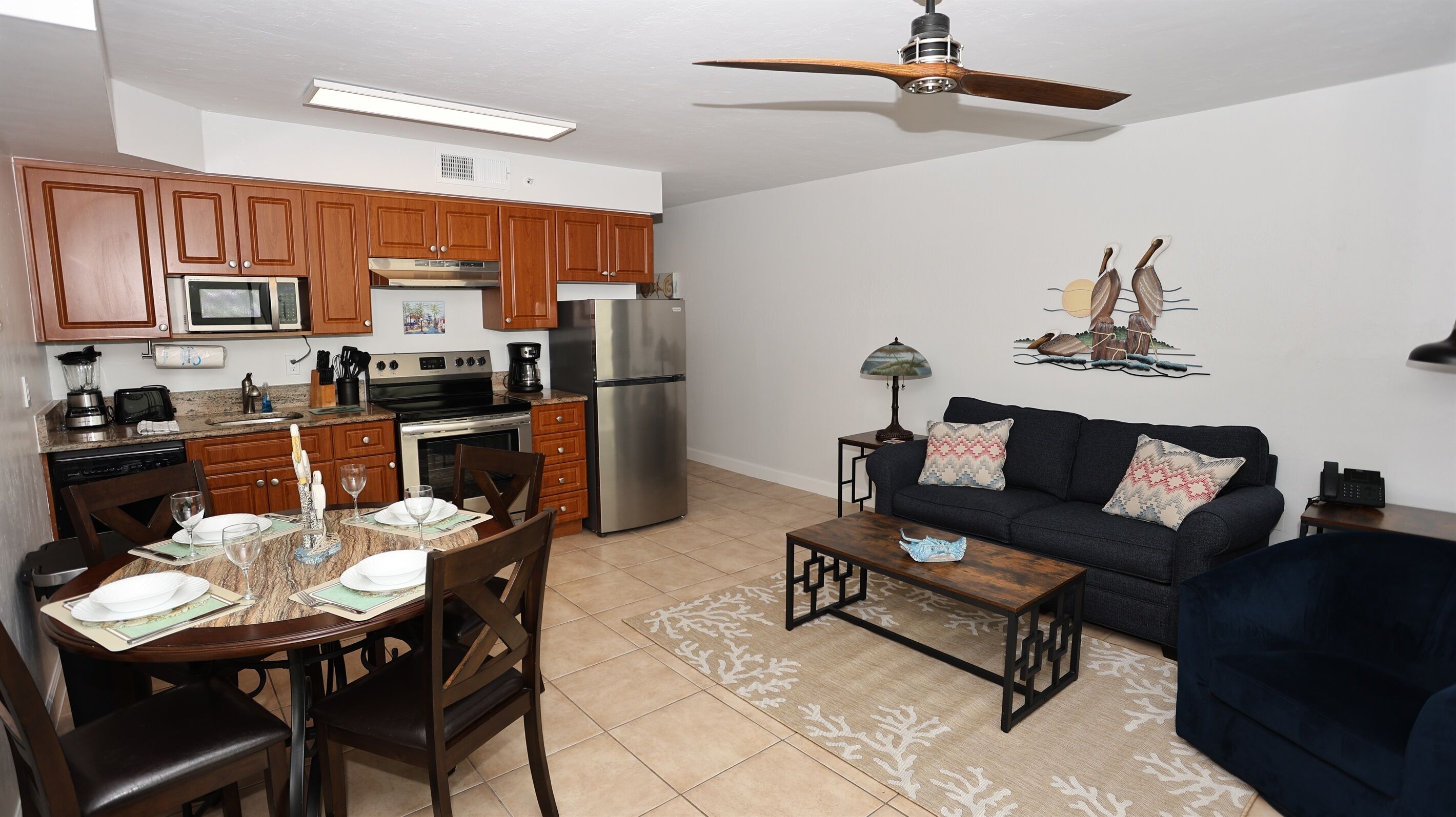 A modern kitchen with wooden cabinets, a refrigerator, stove, and microwave. There is a dining area with a table set for two, chairs, and a coffee machine. A living area with a sofa and a lamp is visible.