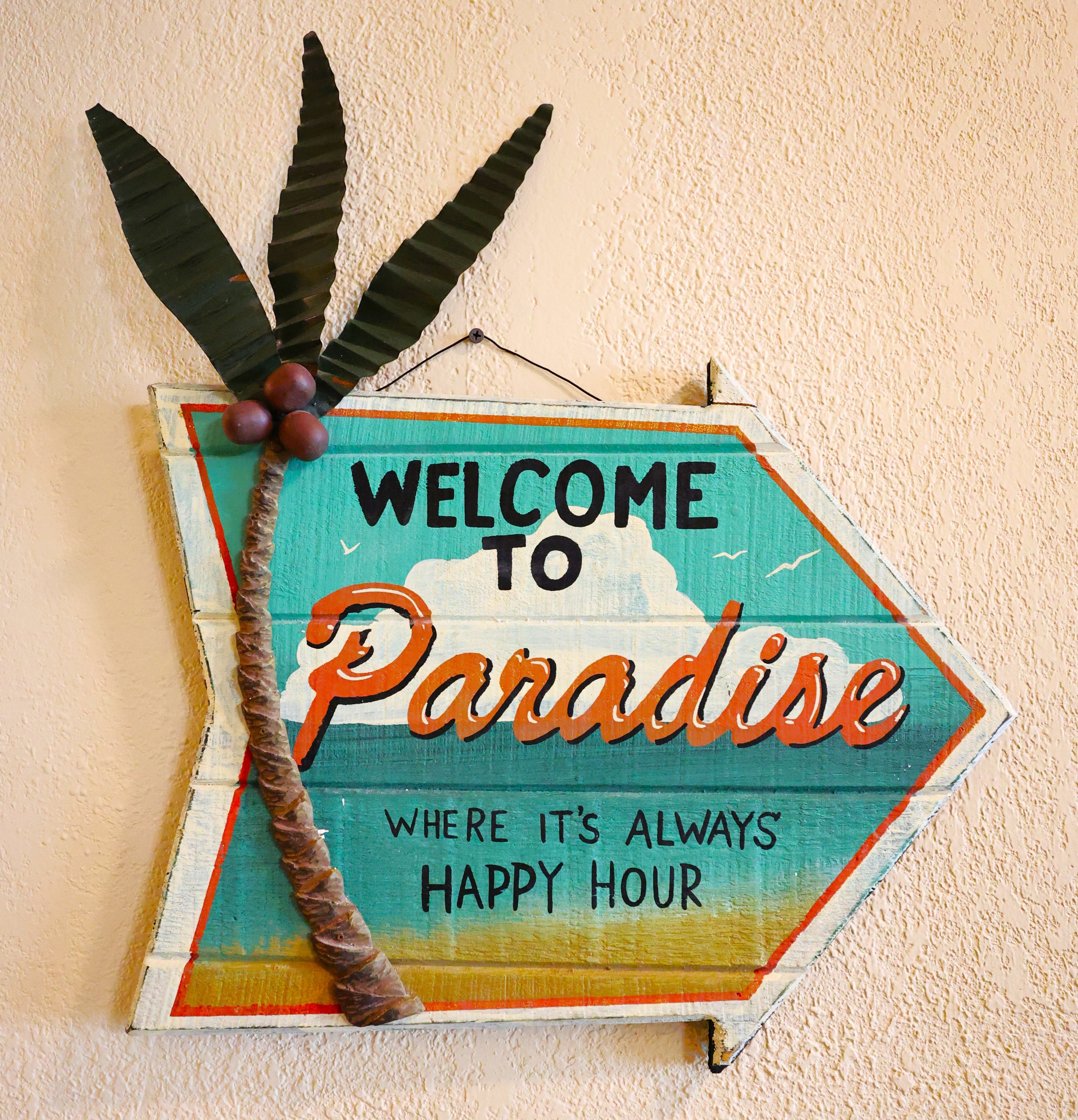 Ein Willkommensschild mit dem Schriftzug 'WELCOME TO Paradise' und 'WHERE IT'S ALWAYS HAPPY HOUR' vor einer strukturierten Wand.