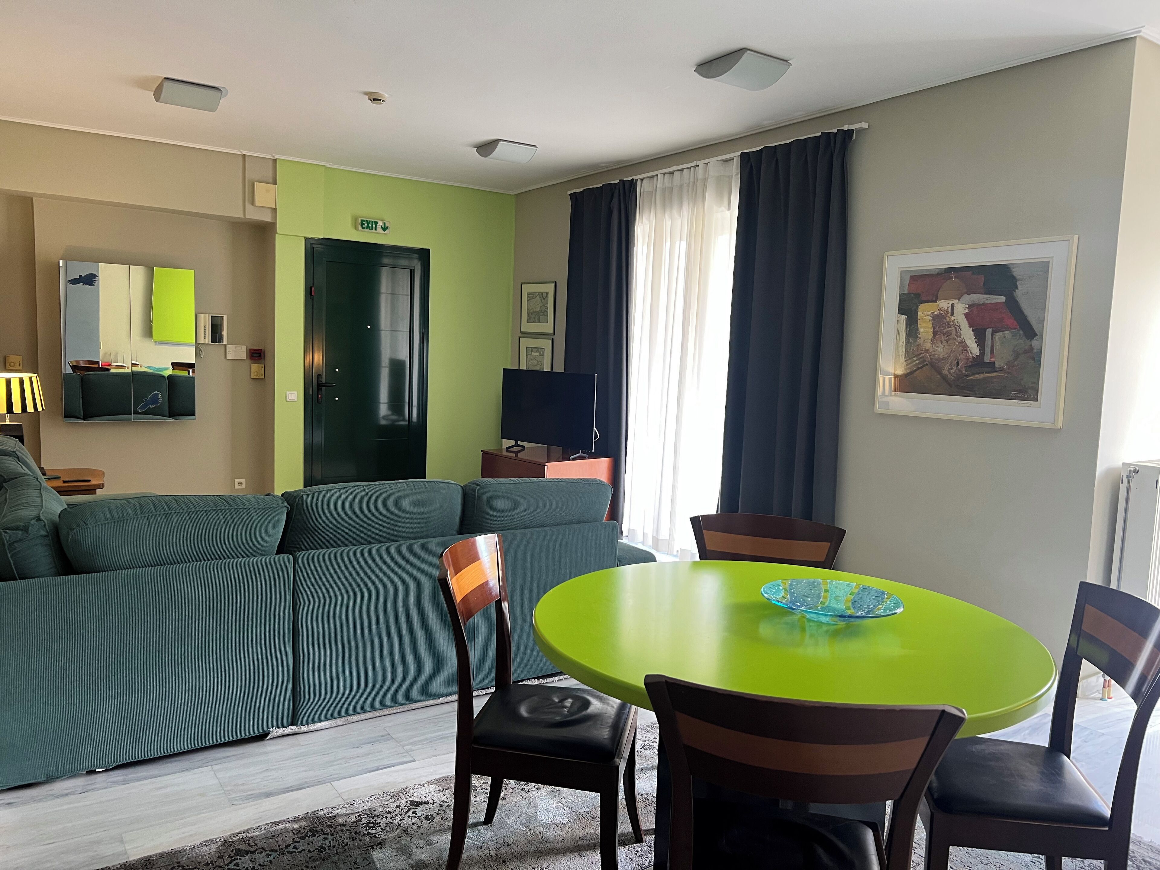 A modern living room with a green sofa, a round green table with chairs, a flat-screen TV, and a framed abstract painting on the wall.