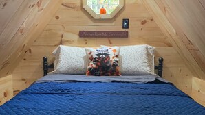 A cozy attic bedroom with a wooden sloped ceiling, a quilted blue bedspread, and a raccoon-themed pillow. - Raccoon Lodge at Coops Creek Cabins (Dunlap)