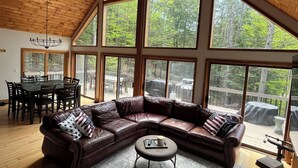 A spacious living room with a large L-shaped sectional sofa, a dining table with chairs, and large windows offering a view of the outdoors. - Modern Chalet Near Mt. Sunapee & Lake Massasecum (Bradford)