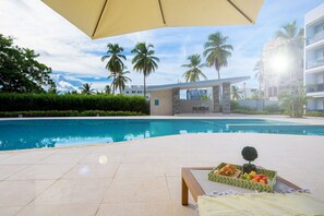 A poolside area with a lounge chair, a table with a fruit tray, and a view of a modern building and palm trees.