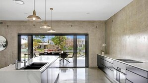 A modern kitchen with a large island, stainless steel appliances, pendant lighting, and a view through glass doors. - Luxico Urban Hideaway (Pet Friendly) (Broadbeach Waters)