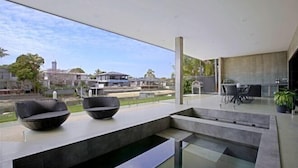 A modern outdoor seating area with a glass table and chairs, a concrete bench, and a view of a residential area with houses and trees. - Luxico Urban Hideaway (Pet Friendly) (Broadbeach Waters)